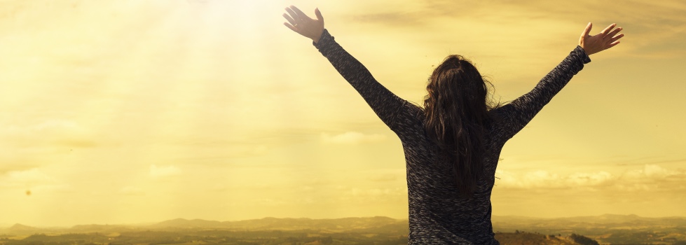 A woman stands facing the horizon and golden sky with her arms raised wide above her head 